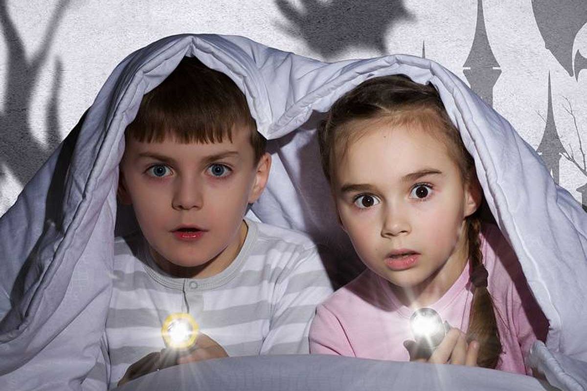 Duas crianças pequenas em baixo do edredom, segurando pequenas lanternas acessas, ilustram o nosso artigo sobre: Como ensinar as crianças a lidar com o medo.
