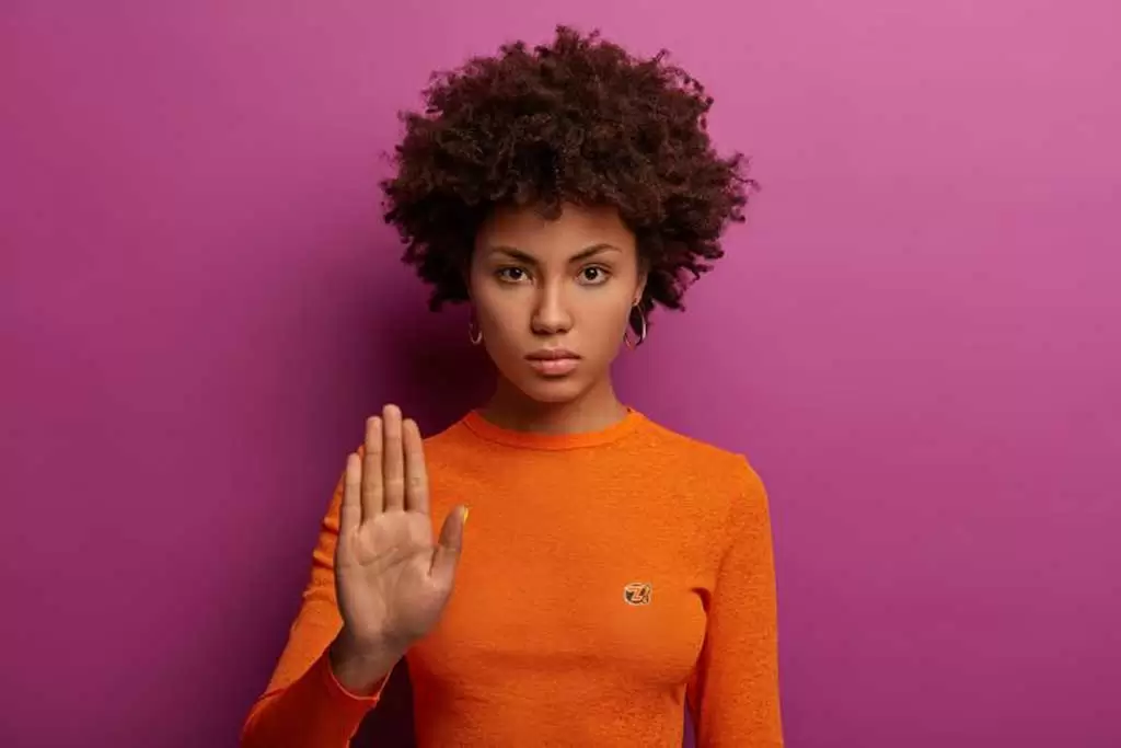 Mulher séria faz gesto de pare, veste suéter laranja isolado na parede roxa ilustra nosso artigo sobre: Qual a importância de estabelecer limites?