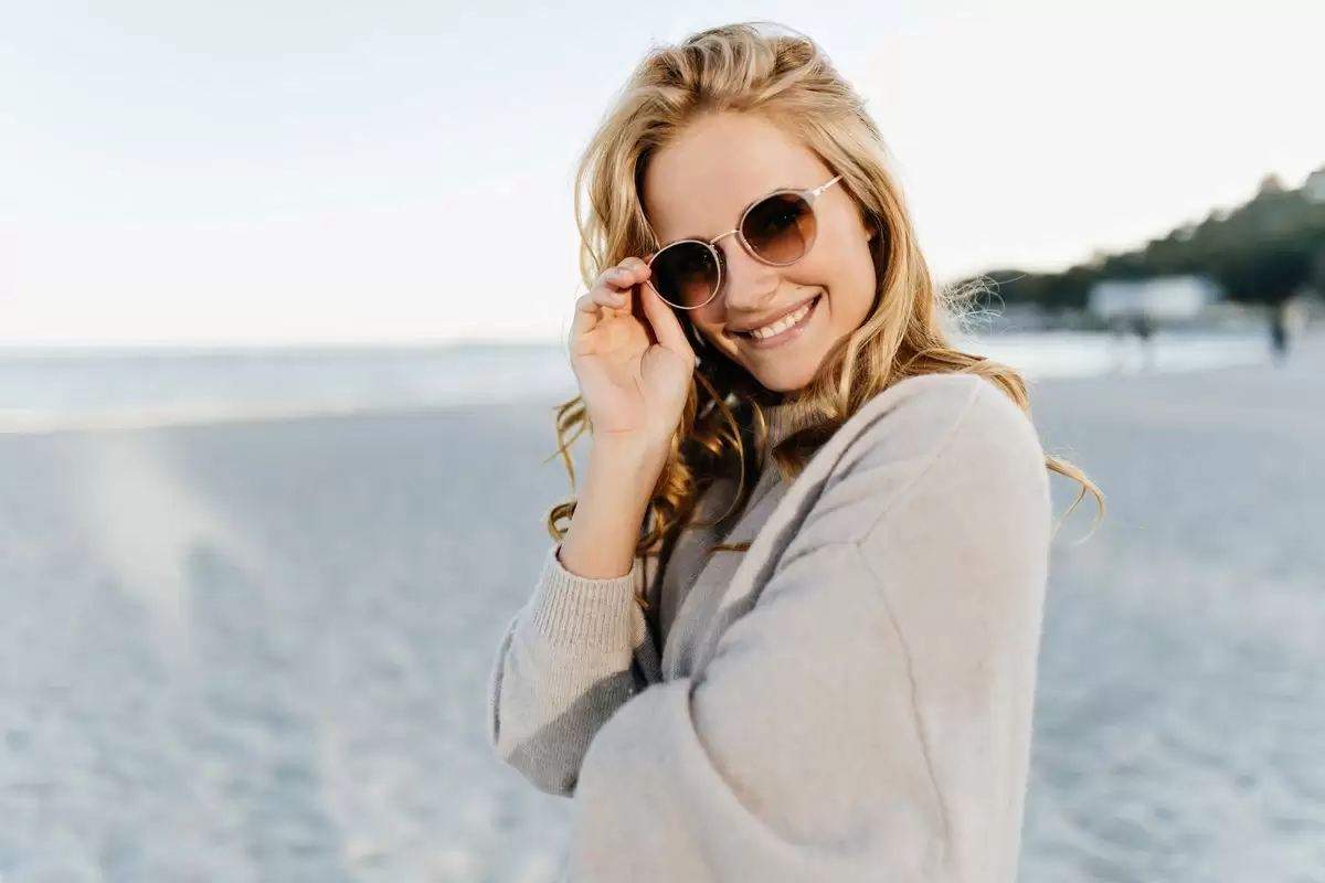 Uma mulher usando óculos escuros posa para foto em frente as ondas da praia ilustrando nosso artigo sobre: Por que é importante ressignificar nossas experiências.