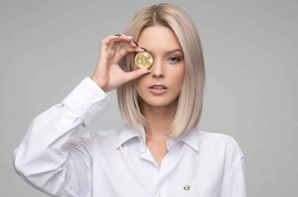 Mulher loira jovem segurando uma moeda de bitcoin em frente ao rosto