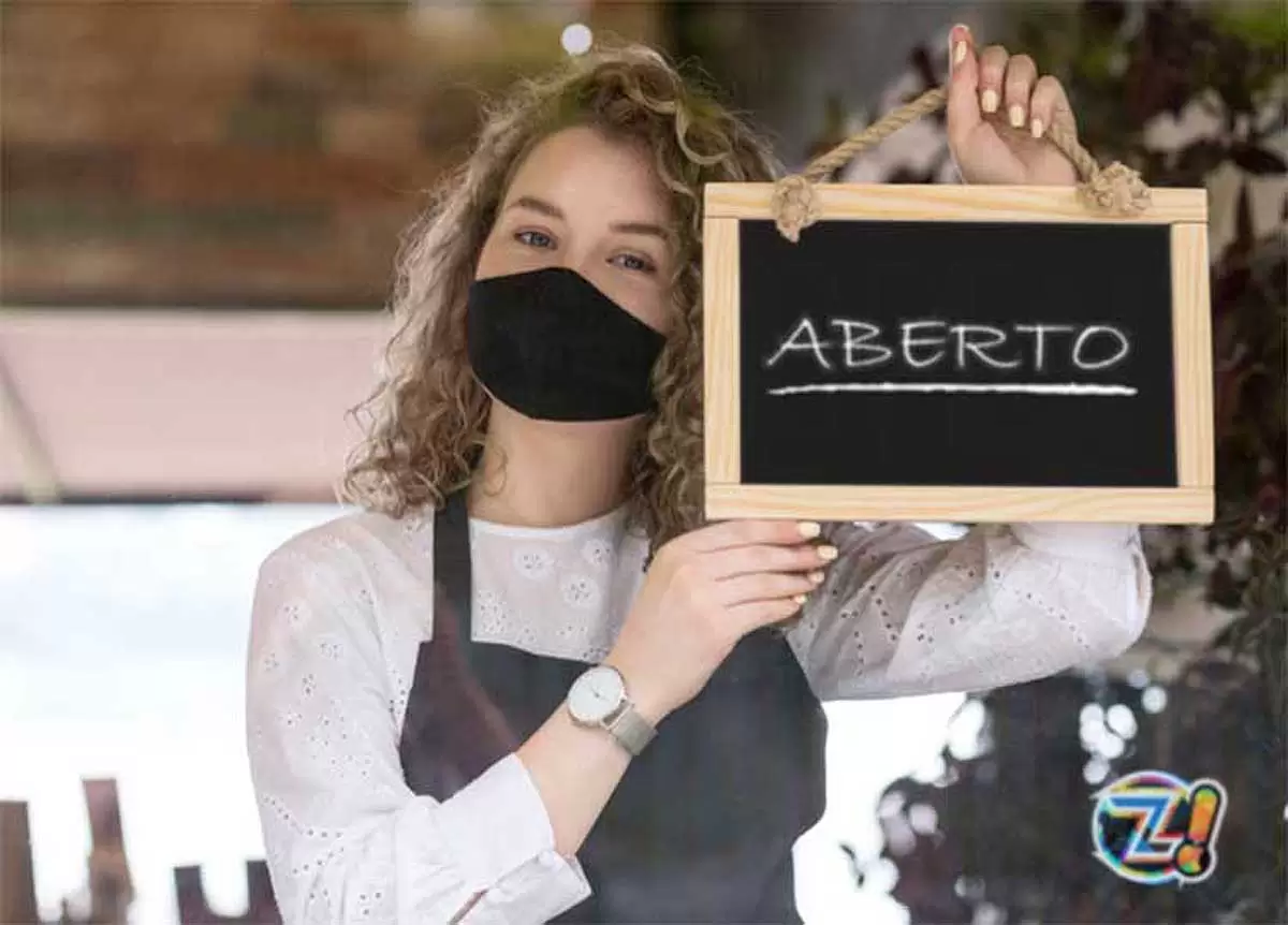 Uma mulher jovem usando máscara e segurando uma plaquinha com a palavra: aberto, ilustra nosso artigo sobre: Como abrir uma empresa em São Paulo Uma mulher jovem usando máscara e segurando uma plaquinha com a palavra: aberto, ilustra nosso artigo sobre: Como abrir uma empresa em São Paulo