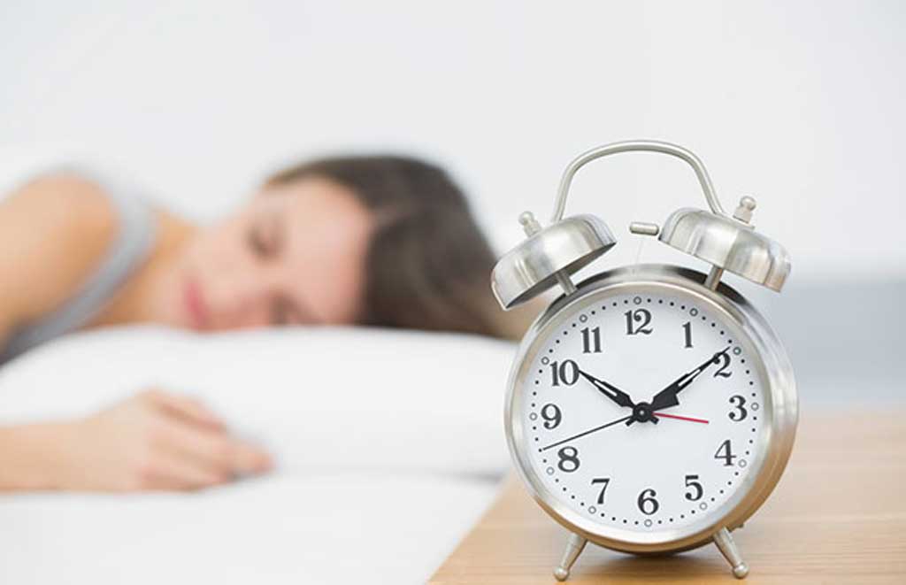 Um relógio despertador ao lado da cama onde há uma mulher domirndo ilustra nosso artigo sobre: Sonhou com relógio.