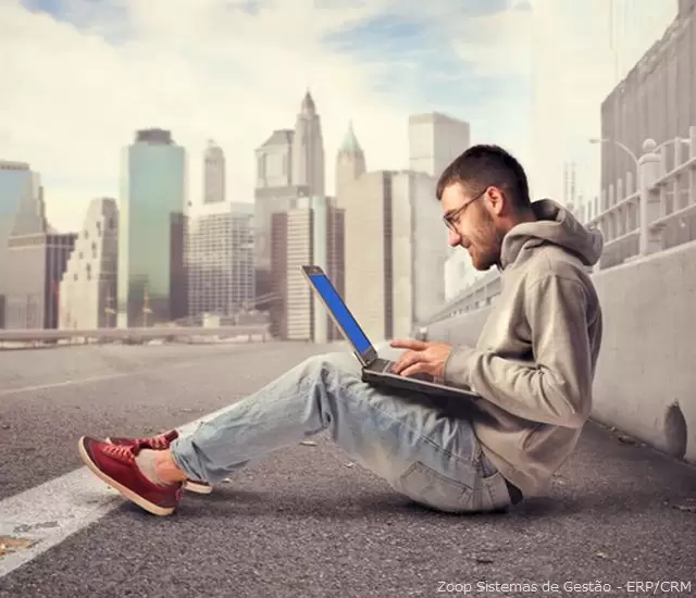 Um homem sentando a beira de uma rodovia usando um notebook tendo ao fundo prédios altos de uma grande cidade ilustra nosso artigo sobre: Como usar a busca social do Facebook.