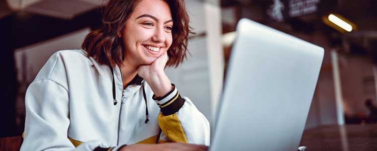 Uma mulher jovem sorridente olhando para a tela de seu notebook ilustra nosso artigo sobre: Veja 9 aplicativos para ganhar uma renda extra.