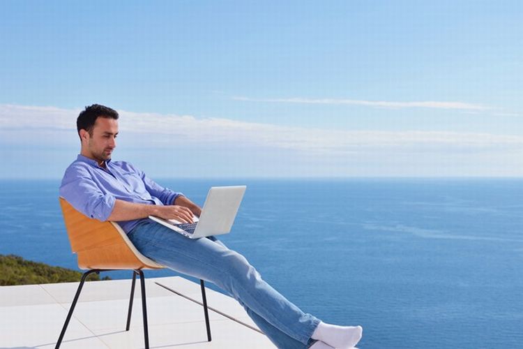 Um homem jovem sentando numa cadeira alaranjada trabalha em seu notebook tendo o mar mediterrâneo ao fundo, ilustra nosso artigo sobre: trabalho freelance. 
