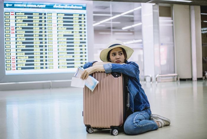 Uma mulher jovem sentada no chão abraçada a sua mala de viagem no saguão de uma aéroporto.