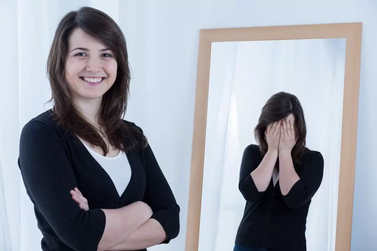Uma mulher sorri olhando para a câmera enquanto seu reflexo no espelho domonstra expressão de vergonha ilustra nosso artigo sobre: Somos verdadeiras fábricas de boas oportunidades.