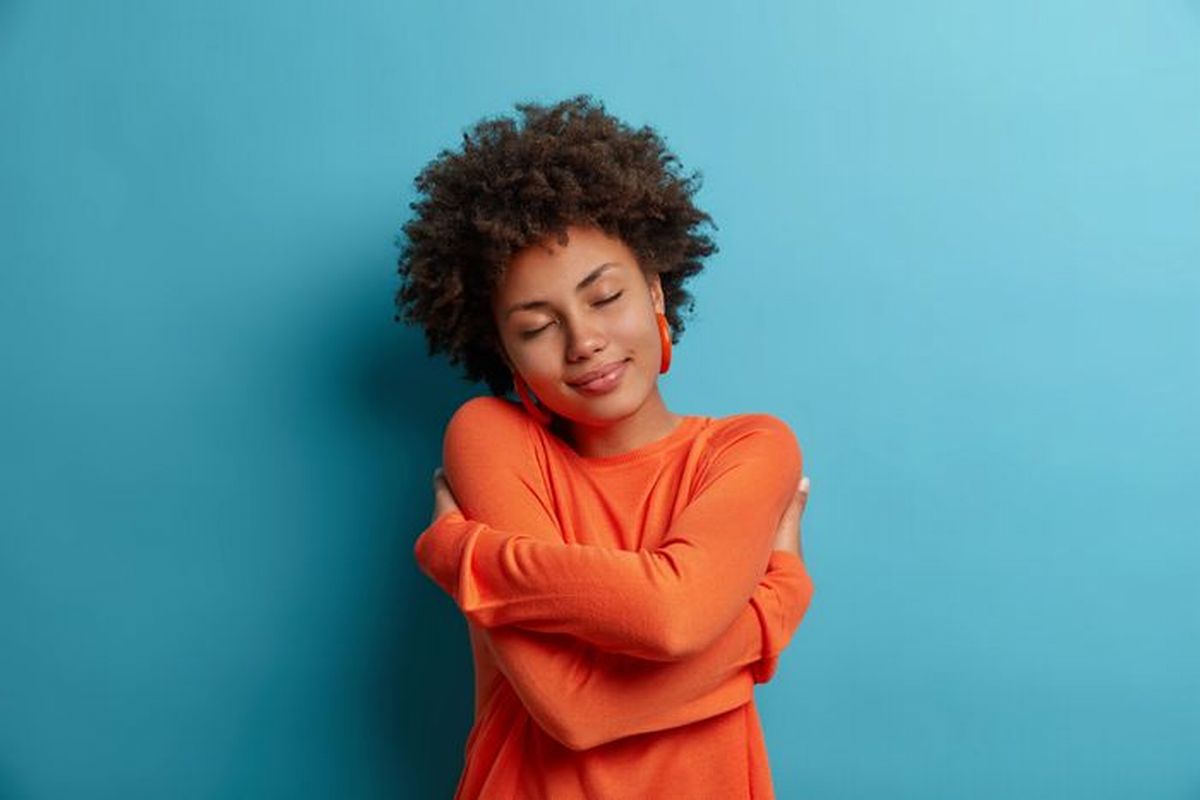 Uma mulher jovem vestindo uma camiseta de cor abóbora envolve o próprio corpo como que num auto abraço ilustra nosso artigo sobre: O que é autocompaixão?
