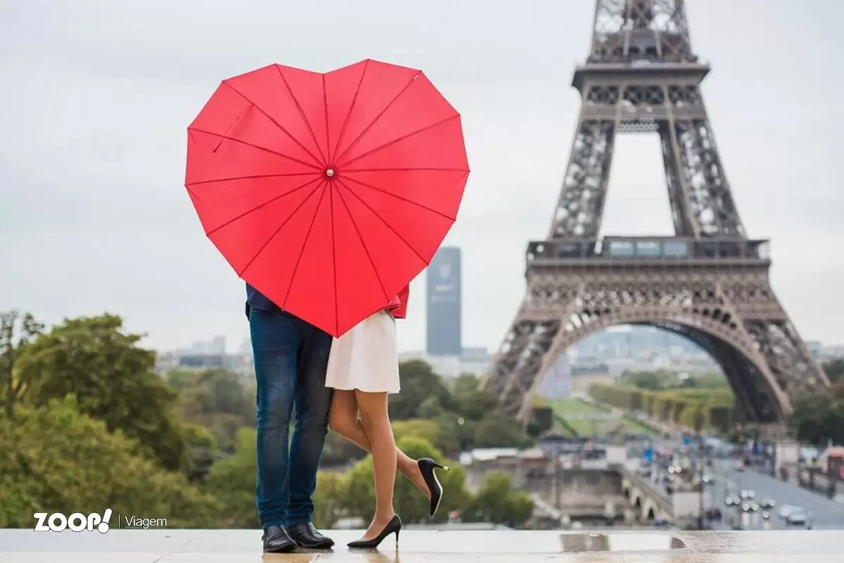 Uma casal se beijando atrás de um guarda-chuva vermelho e em frente a torre Eifel ilustra nosso artigo sobre: Lua de mel: conheça alguns destinos perfeitos para passear.
