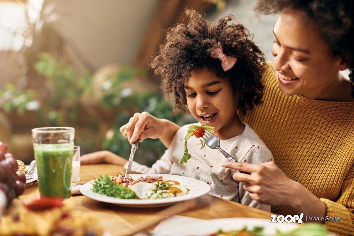 Uma mãe com sua filha no colo durante a refeição ilustra nosso artigo sobre: O que é alimentação saudável e quais seus benefícios. 