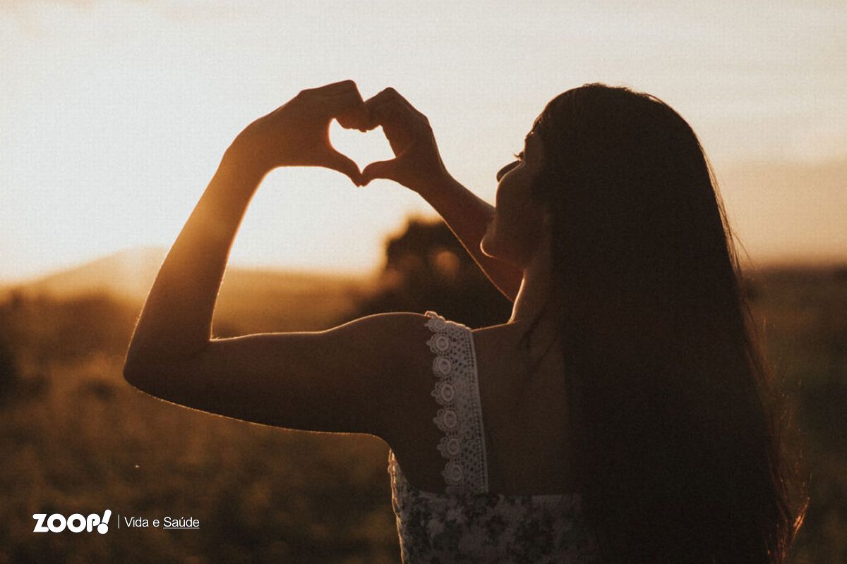 Uma mulher faz um coração com as mãos ao nascer do sol para ilustrar nosso artigo sobre: Autocompaixão.