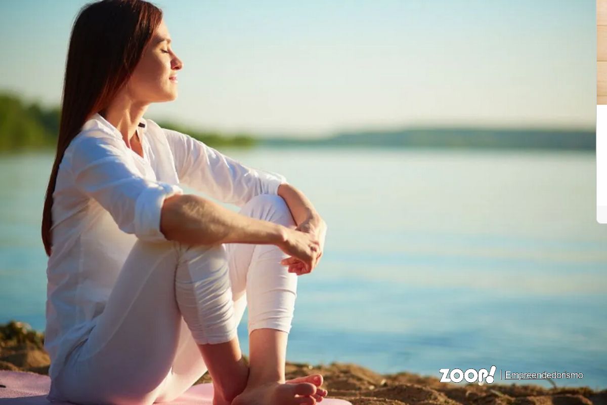 Uuma mulher meditando num dia de sol em frente a um lago ilustra nosso artigo sobre: Autocuidado: conheça a importância para a saúde mental. 